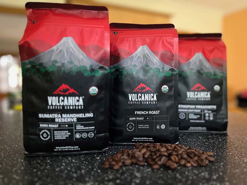 Three bags of Volcanica coffee, their French Roast is in the middle and in focus, some coffee beans on the table in front