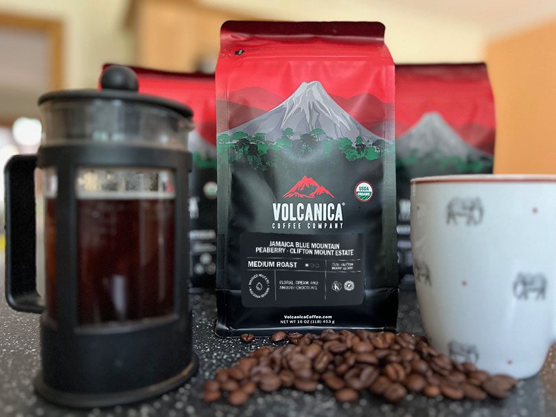 Freshly made French press coffee and a bag of Volcanica Jamaica Blue Mountain Peaberry with some coffee beans spilled in front